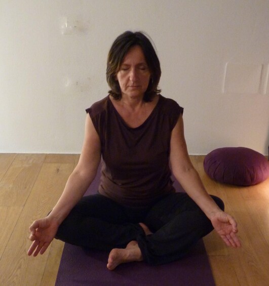 A person sitting cross-legged on a purple mat in a room with wooden flooring and a white wall. 3