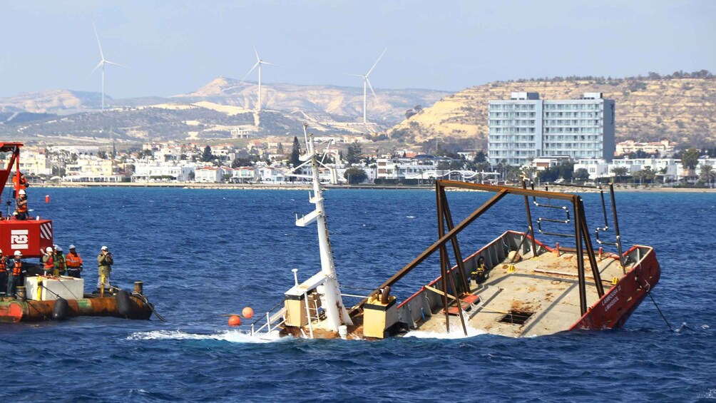 A damaged ship in the water with a crane and workers nearby, a coastal town with buildings and wind turbines in the background. 5