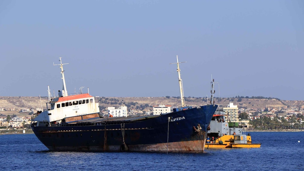 A large ship named 'EL PIDA' docked at a port with a small tugboat nearby, set against a backdrop of a coastal town and hills. 1