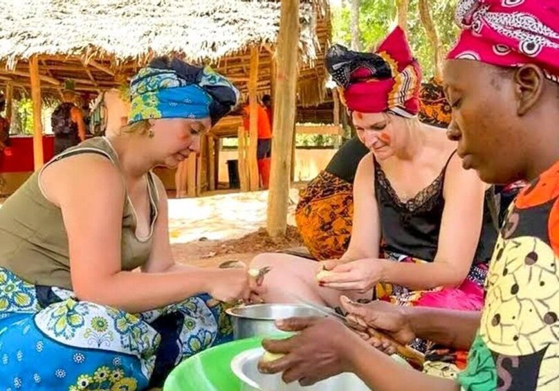 People sitting outdoors, engaged in a communal activity, possibly preparing food, with a thatched roof structure in the background. 1