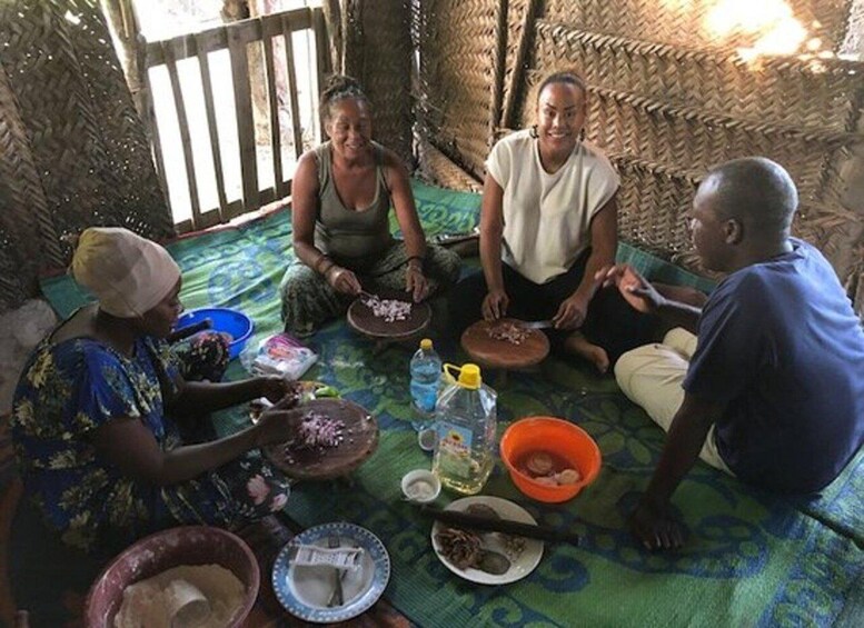 A group of people sitting on a mat inside a hut with woven walls, engaged in a meal with bowls and plates of food. 3