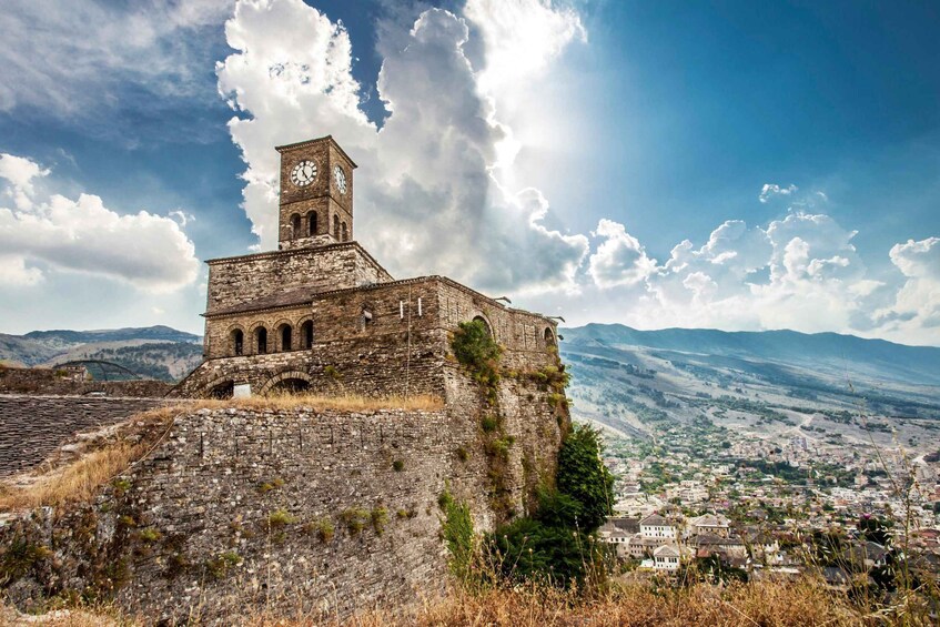 A historic stone building with a clock tower, surrounded by a mountainous landscape and a town below. 5