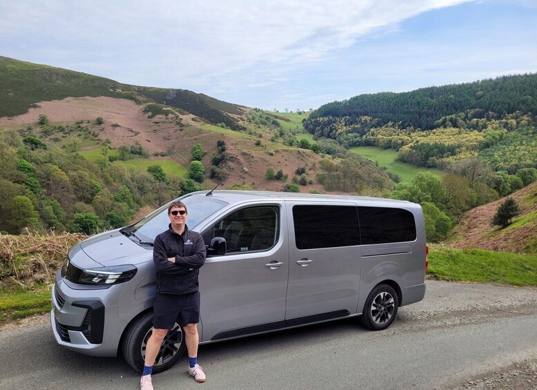 A person standing next to a silver van on a road with a hilly landscape in the background. 1