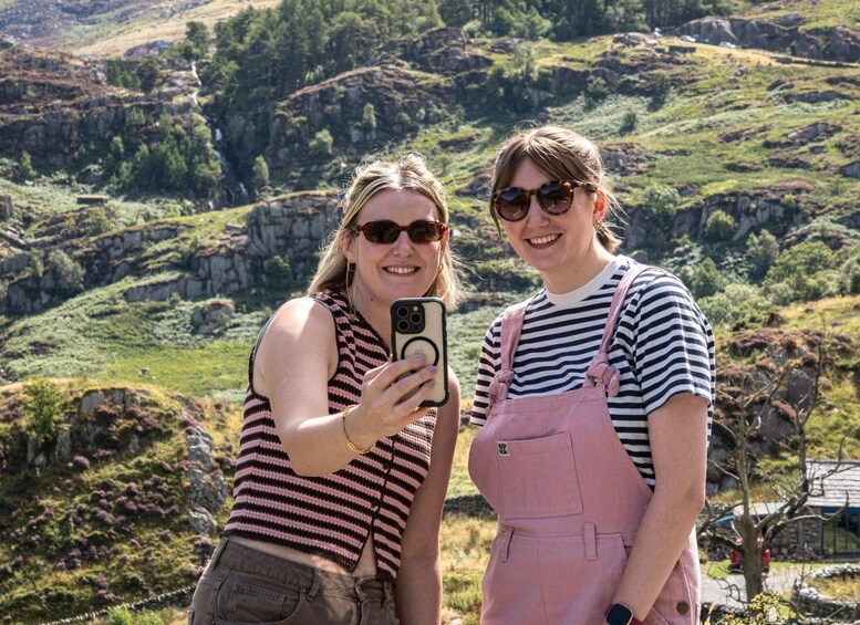 Two people taking a selfie with a smartphone, standing in a hilly landscape with greenery and rocky terrain. 1
