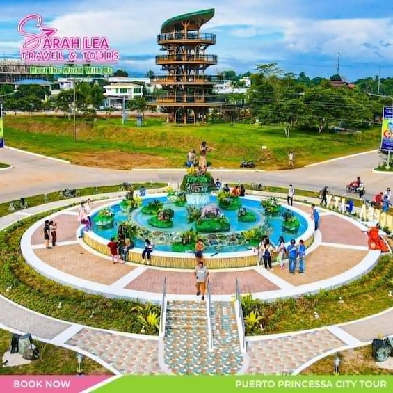 A circular water feature with a central statue, surrounded by a plaza with people and a multi-level building in the background. 2