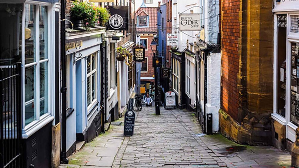 A narrow cobblestone street lined with historic buildings, including a shop with a sign reading 'Bristol Cider Shop.' 3