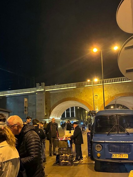 A nighttime street scene with people gathered around a food cart, a blue vintage car, and a bridge in the background. 2