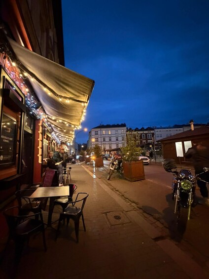 A street at dusk with outdoor seating, string lights, and parked motorcycles. 1