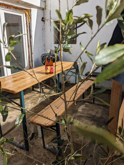 A wooden bench and table in a rustic outdoor setting with a grill and bottles of beer. 4