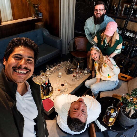 A group of people seated around a table with wine glasses and bottles, in a room with a blue couch and a wine rack in the background. 4