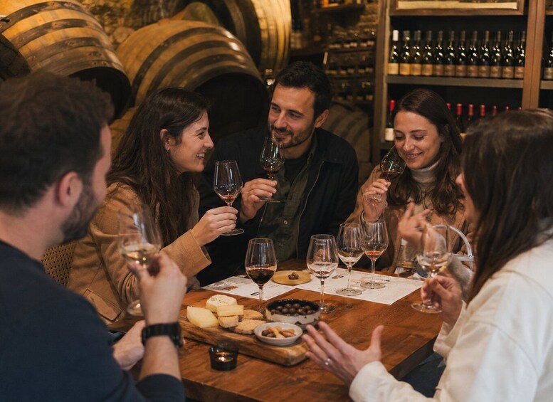 A group of people toasting with wine glasses at a wooden table with cheese and olives, in a setting with wine barrels and shelves of wine bottles. 3