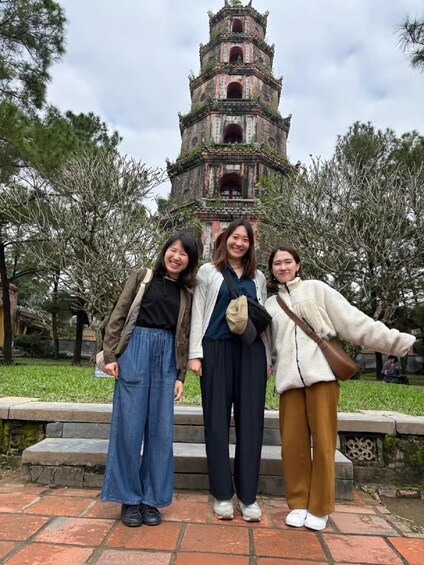 Three people standing in front of a historic tower with multiple levels and a staircase leading up to it. 1