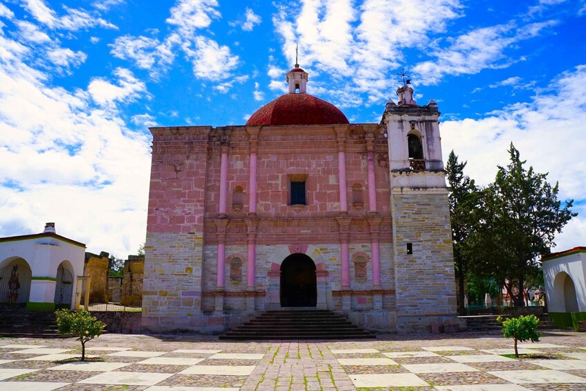 A historic building with a red dome and bell tower, surrounded by a paved courtyard with trees. 1