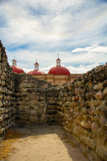 A stone wall with a narrow passage leading to a building with red domes and a bell tower. 2