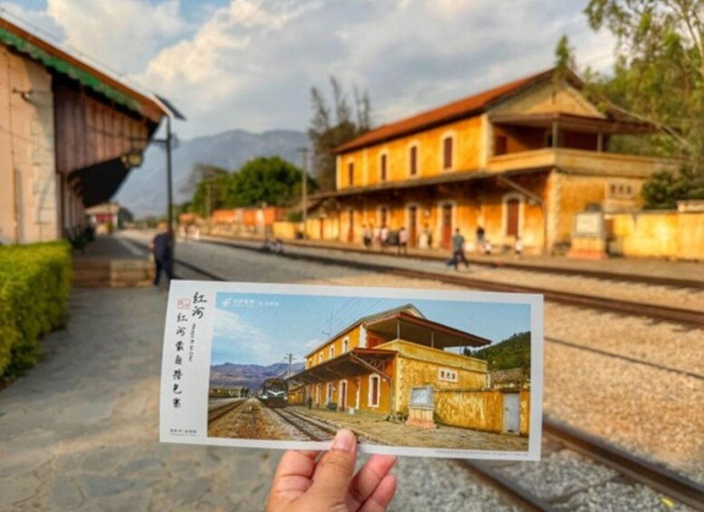 A hand holding a postcard with a photo of a train station and a building with a red roof. 1