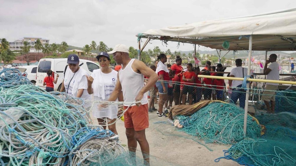 People standing near a pile of fishing nets and ropes, with a tent and parked cars in the background. 2