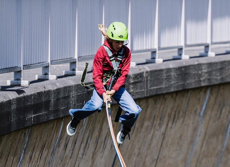 A person in a red jacket and green helmet rappelling down a concrete structure. 1