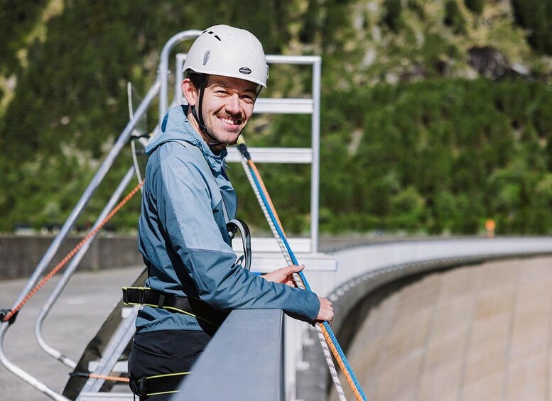 A person in a blue jacket and helmet, holding a measuring tape, standing on a structure with a metal ladder and railings, with greenery in the background. 5