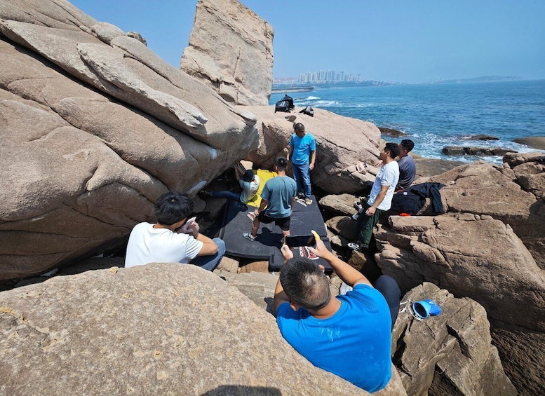 A group of people sitting and standing on rocky terrain by the sea, with a city skyline in the background. 2