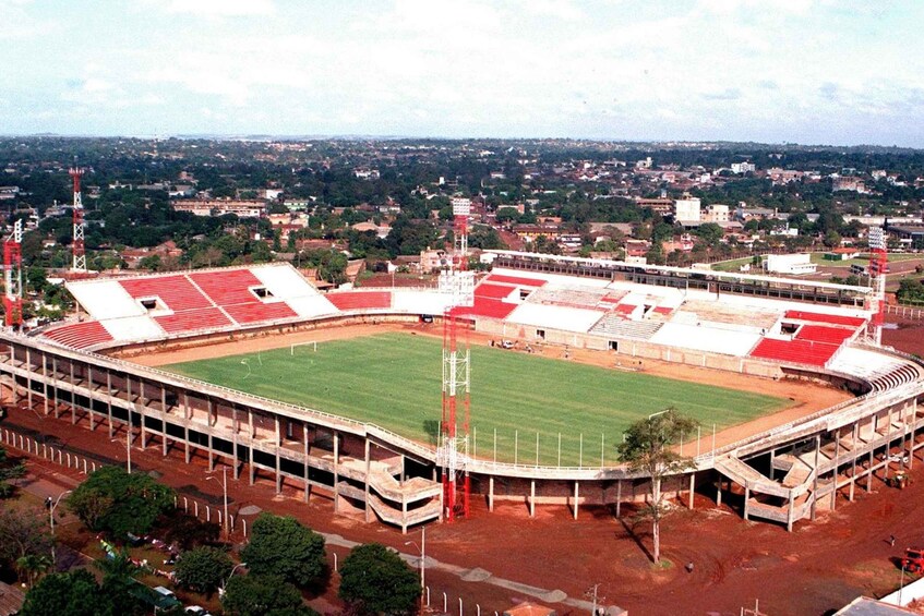 A large stadium with a green field and red seating areas, surrounded by trees and buildings. 2