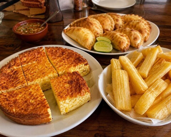 A plate of empanadas, a plate of fried plantains, and a slice of cake on a wooden table. 2