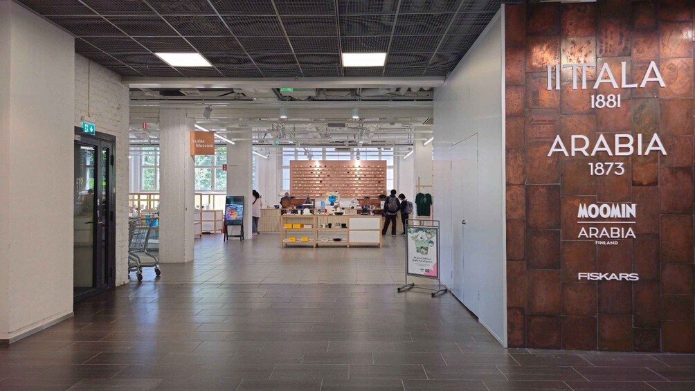 An interior space with a display of items, a sign reading 'ITALA 1881 ARABIA 1873 MOOMIN ARABIA FINLAND FISKARS,' and a tiled floor. 1