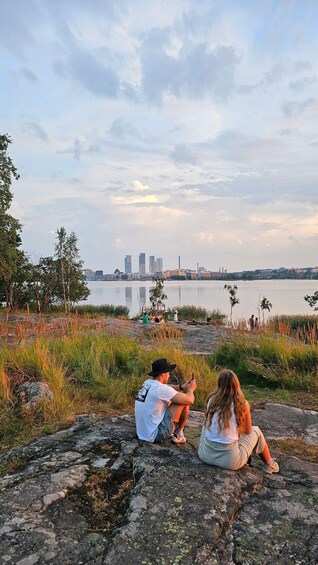 Two people sitting on a rocky shore by a calm lake with a city skyline in the background. 2