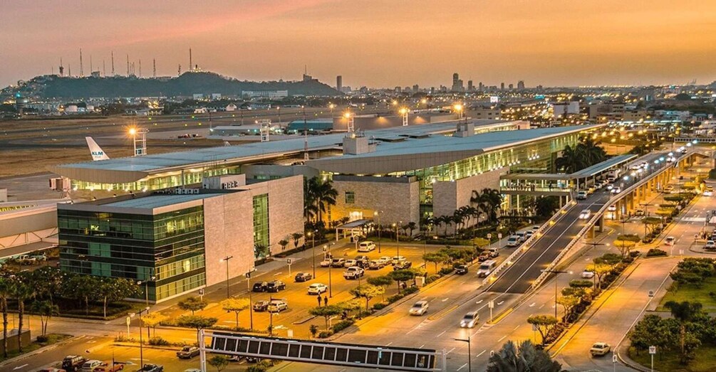 An airport terminal with a modern design, surrounded by roads and a city skyline at dusk. 1