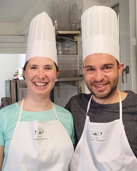 Two people wearing chef hats and aprons with logos, standing in a kitchen. 3