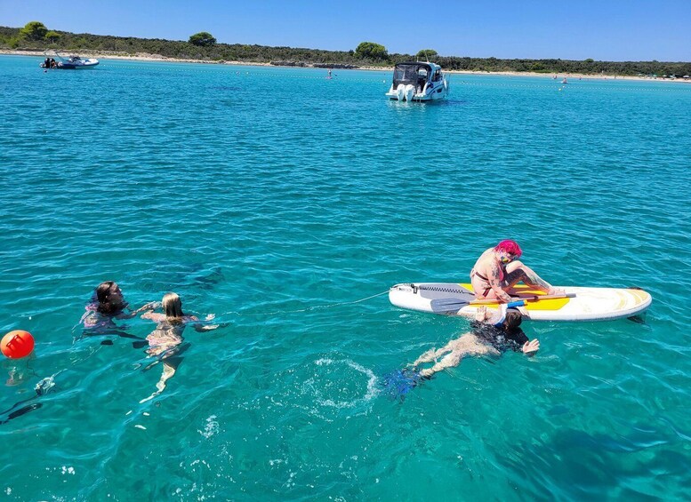 A group of people swimming and paddleboarding in clear turquoise waters with a beach and boats in the background. 2