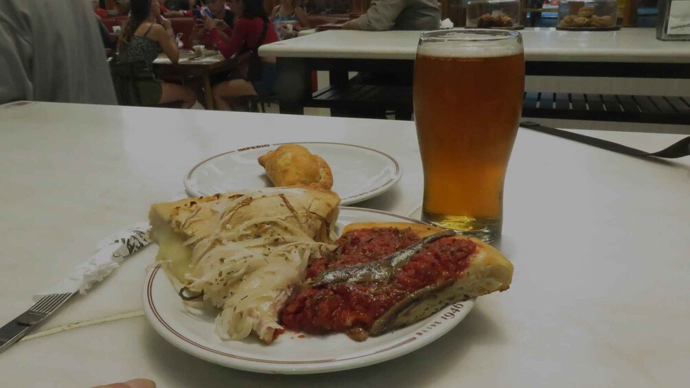 A plate with a slice of pizza and a piece of bread with tomato sauce, a glass of beer, and a fork on a table. 3