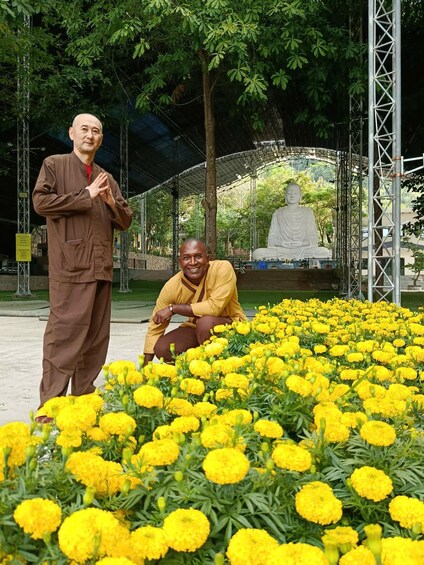 Two individuals in traditional attire, one standing and one kneeling, surrounded by a field of yellow marigold flowers, with a large white statue in the background. 5
