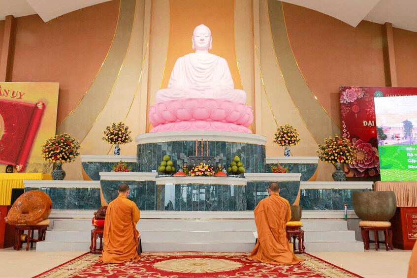 A large statue of a seated figure in white and pink, surrounded by two monks in orange robes, with a red carpet and floral decorations. 1
