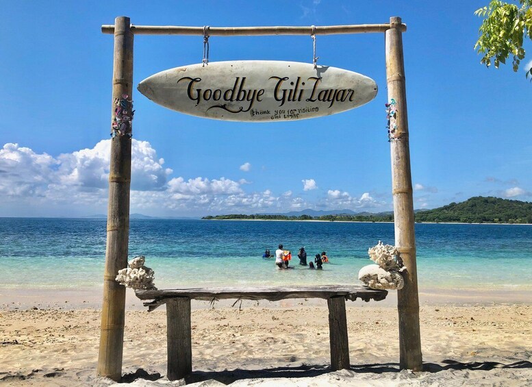 A wooden swing with a sign that reads 'Goodbye Gili Layar' and a beach with clear blue water and distant islands. 5
