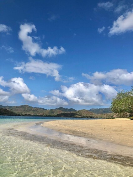 A beach with clear water, sandy shore, and distant mountains under a partly cloudy sky. 1