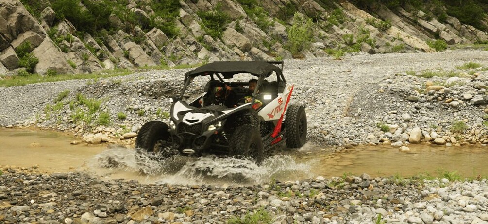 A white and red off-road vehicle crossing a shallow stream in a rocky terrain. 1