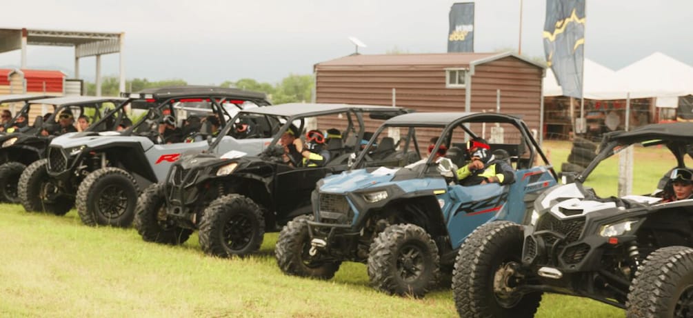 A line of ATVs parked on grass with people inside, a building, and flags in the background. 1