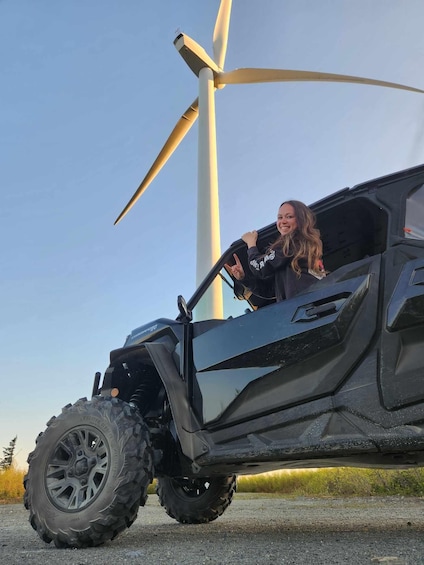 A person in a black vehicle with large off-road tires, parked near a wind turbine. 1