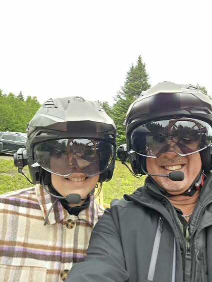 Two people wearing helmets and safety gear, standing outdoors with trees and a car in the background. 4