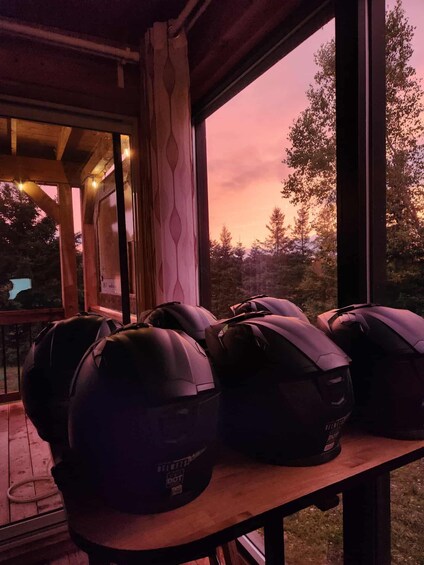 A wooden table with several helmets on it, a window with a view of trees and a sunset. 5