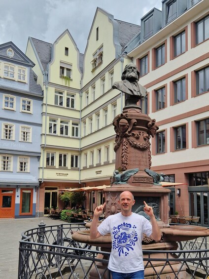 A man standing in front of a statue with a bird and a bust, with colorful buildings in the background. 4