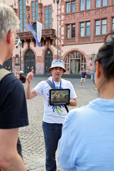 A man holding a tablet with a cityscape image, standing in front of a historic building with arched windows and a clock tower. 2