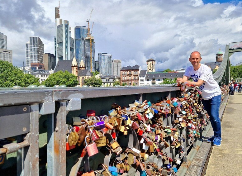 A bridge with numerous padlocks attached to its railing, a city skyline in the background, and a person leaning on the railing. 1