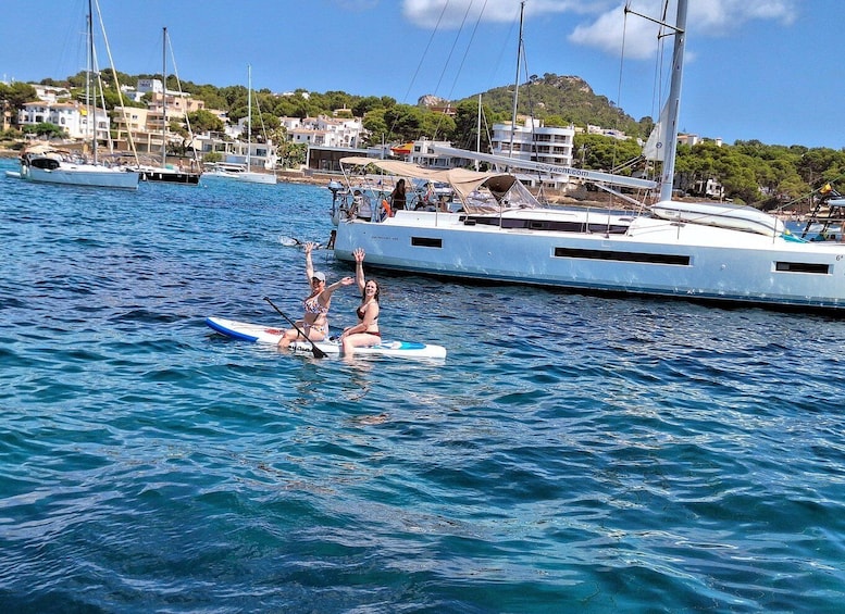 Two people on a paddleboard in calm waters, with sailboats and a coastal town in the background. 4