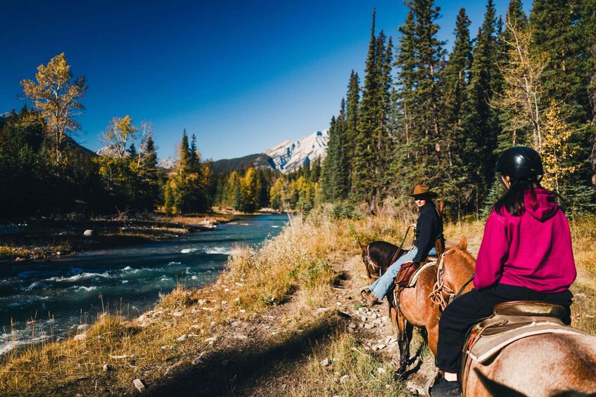 Two people horseback riding beside a river, surrounded by trees and mountains. 1