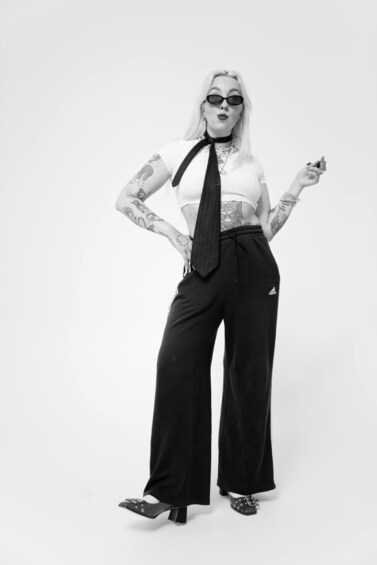 A person with tattoos, wearing a white top, black tie, and black pants, standing against a plain background. 4