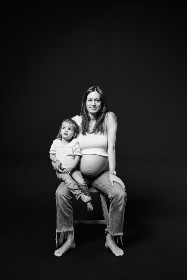 A pregnant woman and a child sitting on a stool against a dark background. 1