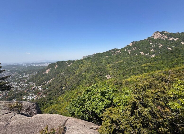 A mountainous landscape with dense green vegetation, a rocky outcrop in the foreground, and a distant cityscape. 4
