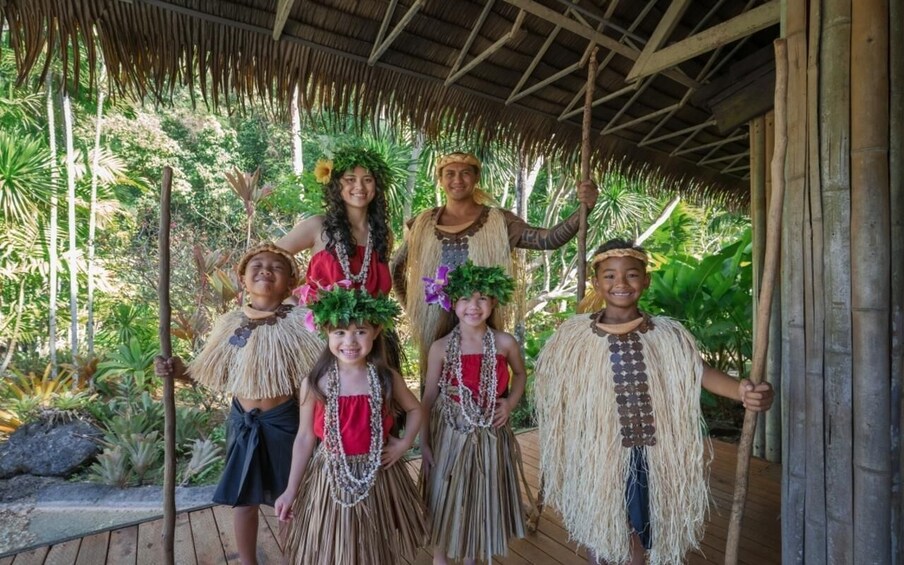 A group of people in traditional attire standing under a thatched roof structure surrounded by tropical vegetation. 4