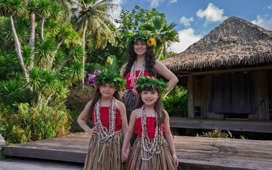Three people in traditional attire with a thatched-roof hut and tropical vegetation in the background. 2